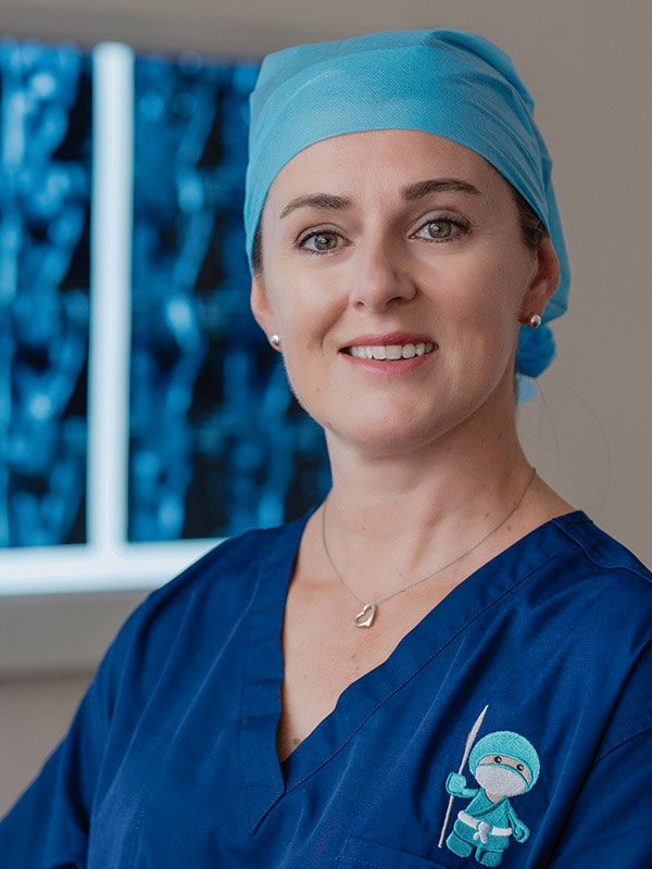 Photo of orthopaedic surgeon Dr Danielle Wadley wearing scrubs standing in front of a background of medical imaging out of focus.